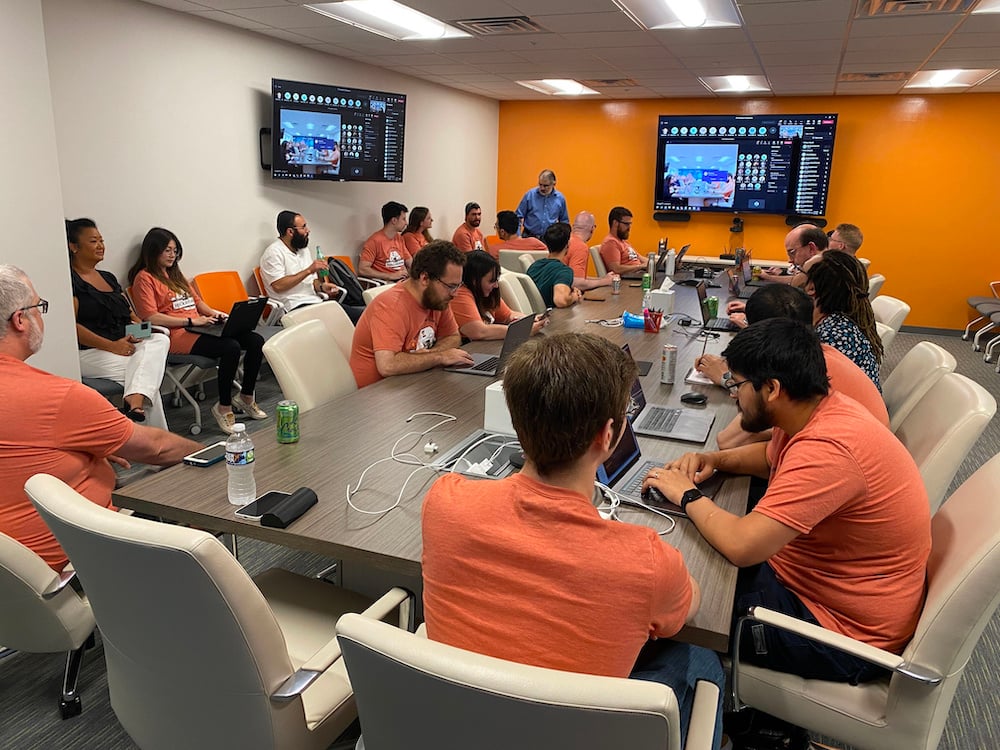 A group of people in a conference room with orange walls, sitting around a large table with laptops and devices. Two screens display a presentation at the front of the room.